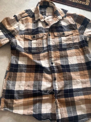 Rutig skjorta från Bluewear, beige/svart - Säljer en rutig skjorta från Bluewear i beige, svart och vitt. Skjortan har långa ärmar, två bröstfickor med knappar och klassisk krage. Tillverkad i mjuk bomull, perfekt för en avslappnad stil.