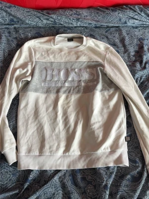 Hugo Boss tröja - Snygg vit sweatshirt från BOSS med stor logga och text i relief på bröstet. Tröjan har rund halsringning, ribbade muddar vid ärmslut och nederkant samt en bred grå panel över bröstet. Perfekt för en clean och stilren look.