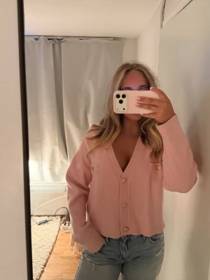 Ljusrosa kofta från Cherry Paris - Supersöt ljusrosa kofta från Cherry Paris i mjuk viskos. Koftan har djup v-ringning, dekorativa fickor framtill och stora guldknappar. Perfekt att styla med jeans eller kjol för en trendig look.