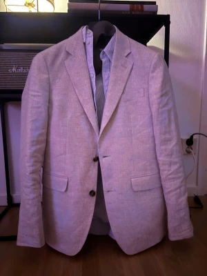 Ljusbeige linnékavaj Dressmann  - Snygg ljusbeige kavaj från Dressmann i 100% linne. Slim fit-modell med två knappar framtill, klassiska slag och två fickor med lock. Perfekt för en stilren och fräsch look. Passar dig som gillar en lätt och luftig känsla.