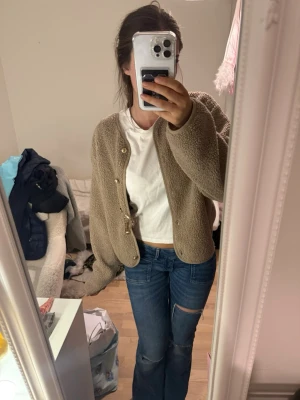 Beige teddy cardigan med knappar - Mysig beige cardiganjacka i teddy-material med rund halsringning och guldfärgade knappar framtill. Jackan har en avslappnad passform och långa ärmar, perfekt för lager på lager under kyliga dagar.