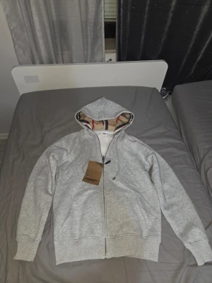 Grå zip-hoodie från Burberry - Snygg grå hoodie från Burberry med dragkedja och klassiskt rutigt foder i huvan. Tröjan har ribbade muddar och är perfekt för en stilren och avslappnad look. Ikonisk Burberry-logga på etiketten och mjukt material för extra komfort.