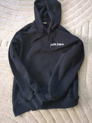 Svart hoodie från Palm Angels - Svart hoodie från Palm Angels med vit logga på bröstet och stor text på ryggen. Klassisk passform med huva och dragsnören samt magficka. Tillverkad i mjukt sweatshirtmaterial, perfekt för en avslappnad streetwear-look. / Insidan har inga Palm angels lappar bara standard