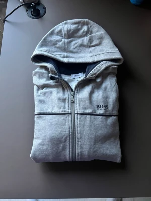 hoodie från Hugo boss  - Snygg ljusgrå hoodie från BOSS med dragkedja framtill och huva. Diskret BOSS-logga på bröstet och mörkare kantband längs fickorna och huvan. Tillverkad i mjukt sweatshirtmaterial, perfekt för en chill och stilren look.