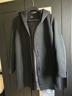 Svart kappa med huva från ONLY - Stilren svart kappa från ONLY med huva och dold dragkedja samt knappar framtill. Kappan har en rak passform och är helfodrad för extra komfort. Perfekt för kyliga dagar och enkel att matcha med olika outfits.