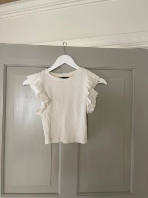 Vit ribbad topp med volangärmar Zara - Supersöt vit ribbad topp från Zara med volangdetaljer på ärmarna. Toppen är ärmlös och har en croppad passform som ger en trendig look. Perfekt att matcha med jeans eller kjol för en fräsch outfit.