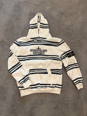 Stone Island Supreme randig hoodie - Vit hoodie med svarta horisontella ränder från Stone Island x Supreme. Stor broderad logga på bröstet och klassisk Stone Island-patch på ärmen. Hoodien har magficka med knappdetalj och ribbade muddar. Perfekt för dig som gillar streetwear och unika samarbeten.