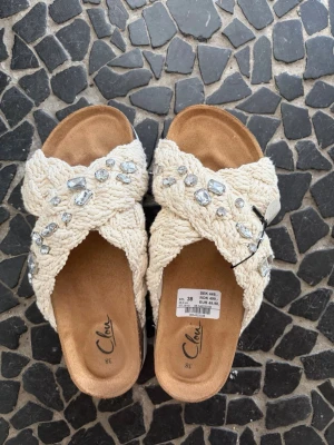 Vita sandaler  - Säljer ett par vita sandaler med bred, flätad ovandel dekorerad med stora glittriga stenar. Sandalerna har öppen tå och en mjuk innersula i beige mockaimitation. Perfekta för en trendig och avslappnad look. Lappen kvar på och inte använda pga försmå
