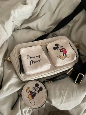  handväska  - Säljer en beige handväska från Disney med två framfickor, båda dekorerade med Mickey Mouse-tryck och text. Väskan har en avtagbar svart axelrem med guldfärgade detaljer och en matchande rund myntbörs med Mickey Mouse-motiv. Tillverkad i konstläder.