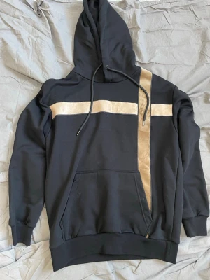 Svart hoodie med guldiga detaljer - Snygg svart hoodie från La Vichy med unika guldiga band i kors över bröstet och längs ena sidan. Klassisk huva med dragsko och stor magficka framtill. Tillverkad i mjukt material som känns skönt mot huden. Perfekt för dig som vill sticka ut lite extra.