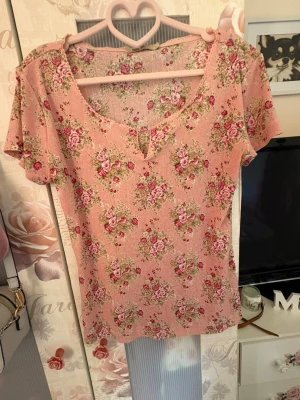 Blommig rosa topp med v-ringning - Supersöt topp i ljusrosa med blommigt mönster i rosa och grönt. Toppen har korta ärmar och en v-ringad hals med liten slits. Perfekt för dig som gillar romantisk och vintage-inspirerad stil. Materialet känns mjukt och lätt.