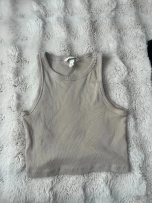Beige ribbad croppad linnetopp - Säljer en beige croppad linnetopp i ribbad bomull. Toppen är ärmlös med breda axelband och rund halsringning. Perfekt till sommaren och enkel att matcha med olika stilar.
