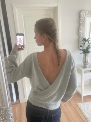 Ljusgrå stickad tröja från NA-KD med öppen rygg - Trendig ljusgrå stickad tröja från NA-KD med djup v-ringning i ryggen och avslappnad passform. Mjuk och fluffig känsla, perfekt till jeans eller kjol. Långa ärmar och ribbade muddar för extra komfort.