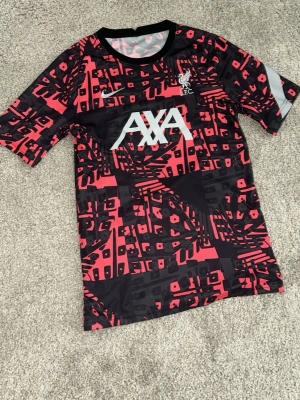 Liverpool Nike Dri-FIT träningströja - Säljer en svart och röd Liverpool FC träningströja från Nike med Dri-FIT material. Tröjan har ett grafiskt mönster, AXA-logga på bröstet och klubbmärke. Perfekt för fotbollsträning eller gymmet.