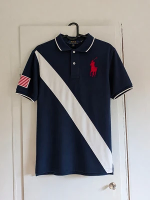 Vintage Ralph Lauren piké - Säljer en snygg och klassisk piké från Polo Ralph Lauren i marinblått. Modellen har en stor vit diagonal rand framtill, röd broderad Polo-logga på bröstet och en USA-flagga på ärmen. Vita detaljer på krage och ärmslut ger ett stilrent och sportigt uttryck. Passar perfekt till jeans, shorts eller chinos. Storlek: L (14-16) Skick: Fint begagnat skick utan defekter! Färg: Marinblå/vit/röd Kan skicka fler bilder eller mått vid intresse!
