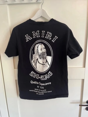Svart Amiri Zig-Zag t-shirt - Cool svart t-shirt från Amiri med stort Zig-Zag tryck på ryggen och mindre logga framtill. Klassisk rund hals och korta ärmar. Tillverkad i mjuk bomull för skön känsla och stilren look. Passar som small/medium