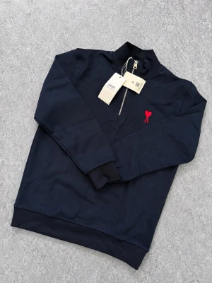 AMI Quarter-zip - Snygg mörkblå tröja från AMI med half-zip dragkedja och hög krage. På bröstet finns ett rött hjärta med bokstaven A broderat. Tillverkad i mjukt bomullsmaterial och har ribbade muddar vid ärmslut och nederkant. Perfekt för en chill och stilren look. Har från storlek S-XXL