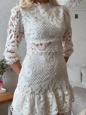 Vit spetsklänning med volang (konfirmationsklänning, studentklänning) - Supersöt vit klänning i spets med trekvartsärm och volangkant nertill. Klänningen har hög hals, genombrutet mönster och markerad midja. Perfekt för dig som vill ha en romantisk och trendig look.