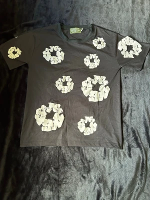 Denim tears T-shirt  - Säljer en svart t-shirt från Denim Tears med stora vita blommönster över hela framsidan. T-shirten har rund hals och korta ärmar. Perfekt för dig som vill sticka ut med en unik och trendig look.