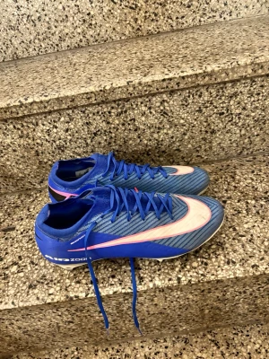 Blåa Nike Mercurial fotbollsskor - Säljer ett par blåa Nike Mercurial fotbollsskor med vit och rosa swoosh på sidan. Skorna har snörning, låg profil och är tillverkade i syntetmaterial för bra grepp och känsla på planen. Perfekta för dig som vill ha snabbhet och kontroll på fotbollsplanen.