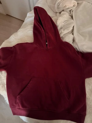 RESERVERAD - Mysig vinröd hoodie med stor huva och klassisk magficka. Hoodien är i mjukt material och har ribbade muddar vid ärmslut och nederkant. Perfekt för chill dagar eller när du vill ha en avslappnad stil. 