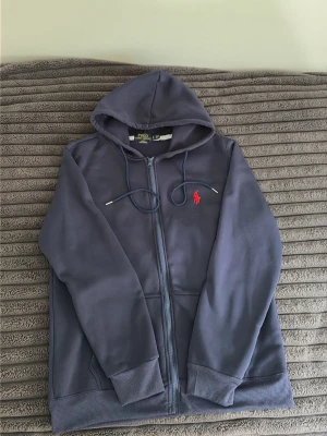 Mörkblå hoodie från Polo Ralph Lauren - Snygg mörkblå hoodie från Polo Ralph Lauren med dragkedja framtill och huva med dragsko. Klassisk röd logga broderad på bröstet. Slim fit-modell i mjukt material, perfekt för en avslappnad stil. Ribbstickade muddar vid ärmar och nederkant.