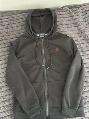 Svart hoodie från Polo Ralph Lauren - Svart hoodie från Polo Ralph Lauren i modellen Slim Fit med stretchmesh-material. Klassisk design med dragkedja framtill och huva. Perfekt för en avslappnad och stilren look.