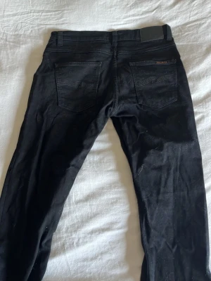Svarta jeans från Nudie Jeans - Säljer ett par svarta jeans från Nudie Jeans med klassisk femficksmodell och raka ben. Jeansen har svarta sömmar och en diskret logga på bakfickan samt svart läderpatch i midjan. Perfekta för en clean och stilren look.