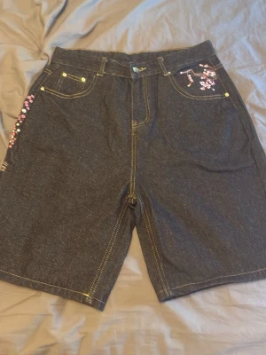 Mörkblå jeansshorts med blommor - Unika mörkblå jeansshorts med broderade rosa och vita blommor på fickorna. Shorts med normal passform, klassiska fickor fram och bak samt kontrastsömmar. Perfekta för dig som vill sticka ut lite extra i sommar.