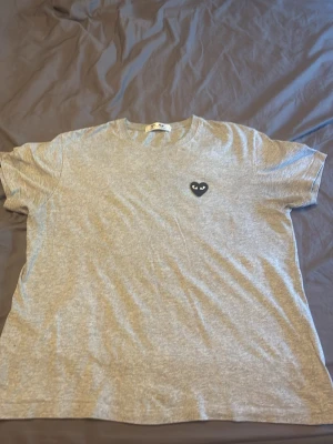 Grå t-shirt med svart hjärtdetalj - Snygg grå t-shirt med rund hals och korta ärmar. På bröstet sitter en svart hjärtformad patch med ögon, vilket ger en unik och cool look. Perfekt till jeans eller shorts för en avslappnad stil.