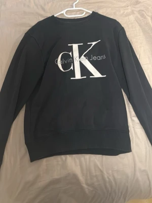 Svart sweatshirt från Calvin Klein - Svart sweatshirt från Calvin Klein Jeans med stort vitt CK-tryck på bröstet. Klassisk rund halsringning och ribbade muddar vid ärmslut och nederkant. Perfekt för en stilren och avslappnad look.