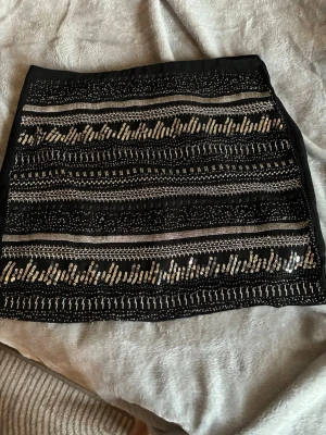 Svart kortkjol med paljetter - Säljer en svart kortkjol med glittriga paljettdetaljer i silver. Kjolen har ett rakt snitt och dragkedja bak. Perfekt för dig som vill sticka ut lite extra med en snygg och trendig look.