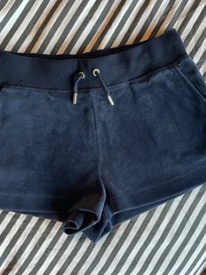 Mörkblå mjuka shorts med snörning - Säljer ett par mörkblå mjuka shorts i från juice couture med snörning i midjan och fickor på sidorna. Shortsen har en broderad detalj vid linningen och är perfekta för sommarens alla chill dagar.
