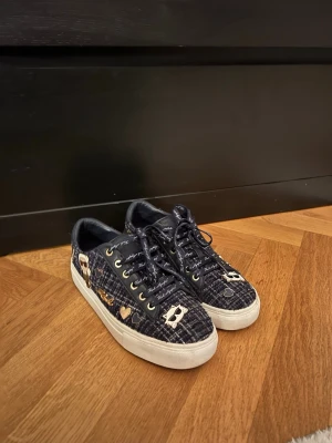 Mörkblå sneakers med guldiga detaljer - Unika mörkblå sneakers med vit sula och guldiga detaljer som hjärtan och figurer. Skorna har snörning, guldfärgade öljetter och ett rutigt tygmaterial som bas. Perfekt för dig som vill sticka ut med coola och lekfulla detaljer.