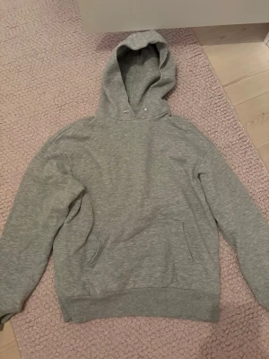 Grå basic hoodie med huva - En enkel och stilren grå hoodie med huva och magficka. Tillverkad i mjukt sweatshirtmaterial, perfekt för chill dagar. Klassisk passform och ribbade muddar vid ärmar och nederkant. Inga synliga loggor eller tryck.