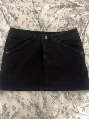 Svart kort jeanskjol med fickor - Säljer en svart kort jeanskjol med fram- och bakfickor samt snygga kopparfärgade knappar. Kjolen har klassisk femficksdesign och är perfekt för dig som gillar en enkel men stilren look.