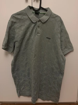 Grå pikétröja från BOSS - Snygg grå pikétröja från BOSS med diskret mönster i tyget och klassisk krage. Tröjan har korta ärmar, knappar vid halsen och BOSS-logga broderad på bröstet. Perfekt för en clean och stilren look.