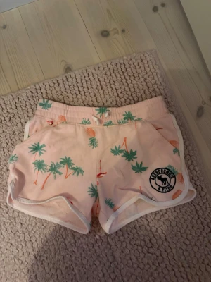 Rosa shorts med palmmönster - Säljer ett par ljusrosa shorts från abercrombie med gröna palmer och orange flamingos. Shortsen har en liten logga på ena benet. Perfekta för sommaren och riktigt sköna att hänga i. Storlek 11/12 år men skulle säga att de är lite större.