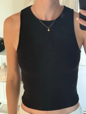 Svart ribbad croppad linnetopp - Snygg svart ribbad linnetopp med croppad passform och öppen rygg. Perfekt för dig som gillar stilrena och trendiga plagg. Toppen är ärmlös och har en tight siluett som framhäver figuren.