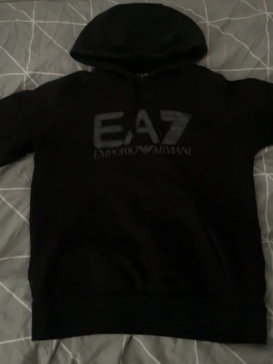 Svart EA7 Emporio Armani hoodie - Snygg svart hoodie från EA7 Emporio Armani med stor logga på bröstet. Klassisk modell med huva och ribbade muddar. Perfekt för dig som gillar stilrena och exklusiva plagg. Materialet känns mjukt och bekvämt.