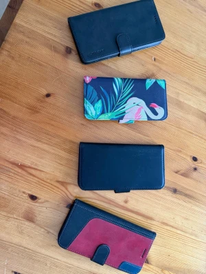 Fyra mobilfodral i olika färger - Säljer fyra mobilskal i plånboksmodell. Tre är svarta, varav ett har vinröda detaljer och ett har ett färgglatt flamingomotiv med gröna blad. Dom passar IPhone 5-7-12, alla  har plats för kort och stängs med magnetlås. Materialet är konstläder och de har öppning för kamera.