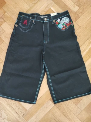 Svarta shorts med broderi och tryck - Unika svarta shorts med färgglada broderier och tryck på fickor och bakstycke. Shortsen har blå kontrastsömmar, knappgylf och bälteshällor. Dekorativa detaljer med blommor och grafiska motiv ger en cool och personlig stil.