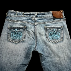 Jeans m. kronor -  Lågmidjade jeans med kronor på bakfickorna. Liknar Victoria Beckham! De är stretchiga. Slitna längst ner. Innerben: 85, midjemått: 84. 
