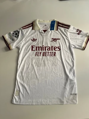 Helt ny! Arsenal tröja 25/26 Eze - Snygg vit Arsenal matchtröja från Adidas med vinröda detaljer och klubbmärke på bröstet. Tröjan har korta ärmar, tryckta sponsorer och nummer 10 med namnet EZE på ryggen. Tillverkad i lätt och ventilerande material, perfekt för fotboll eller träning.