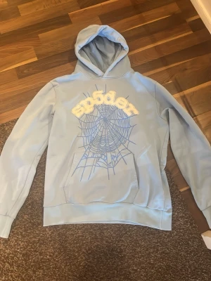 Sp5der, Ljusblå hoodie med spindelnätsmotiv - Sp5der hoodie i storlek M, Säljer en ljusblå hoodie med huva och stor ficka framtill. På bröstet finns ett stort tryck med ett blått spindelnät och texten 'sp5der' i vitt med gulddetaljer. Hoodien har en avslappnad passform och är gjord i mjukt material.