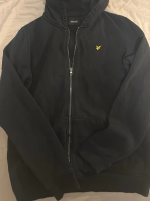 Svart hoodie med dragkedja - Säljer en svart hoodie med dragkedja och huva. Den har en gul broderad logga på bröstet och två fickor framtill. Perfekt för dig som gillar en enkel och stilren look. Materialet känns mjukt och bekvämt.