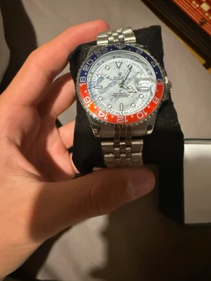  GMT-Master II Pepsi stållänk - Säljer en GMT-Master II med klassisk Pepsi-randig vridring i blått och rött. Klockan har en vit urtavla, datumfönster och silverfärgad stållänk. Perfekt för dig som gillar ikonisk design och vill ha en riktigt snygg accessoar på handleden.