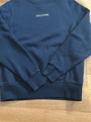 Mörkblå sweatshirt från Hilfiger - Mörkblå sweatshirt med rund hals och diskret Hilfiger-logga broderad på bröstet. Tröjan har långa ärmar, ribbade muddar vid ärmslut och nederkant samt en klassisk, avslappnad passform. Perfekt för en stilren och enkel look.