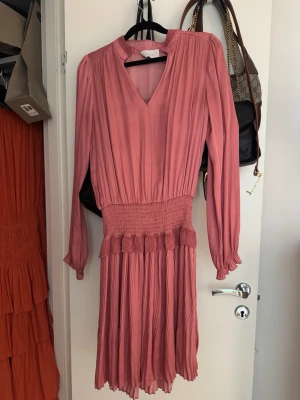 Plisserad rosa klänning med v-ringning - Säljer en plisserad rosa klänning med v-ringning och långärmade ärmar. Klänningen har markerad midja med smock och volangdetalj samt ett mjukt, flytande tyg som ger en snygg siluett. Några små fläckar men tror de går bort i tvätten! 