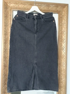 Svart jeanskjol - Rak svart jeanskjol i midi-längd med klassiska fickor fram och bak. Kjolen har en snygg slits framtill och stängs med knapp och dragkedja.